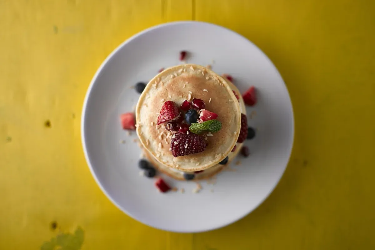 Panquecas empilhadas com frutas vermelhas e coco ralado, em prato branco sobre mesa amarela